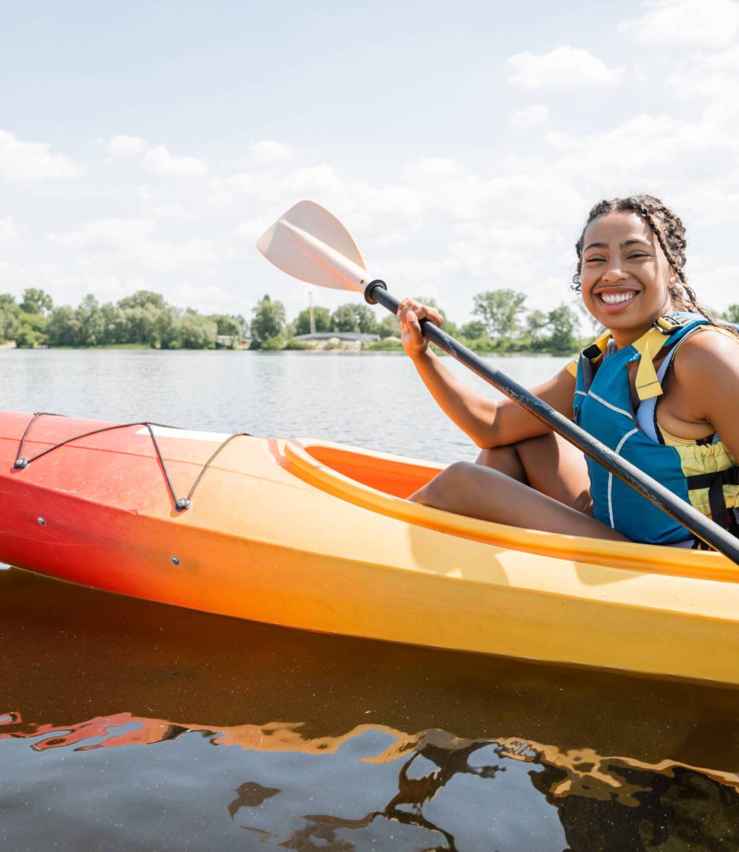 overjoyed african american woman in life vest paddling in sportive kayak and smiling at camera on lake with green picturesque shore on background in summer